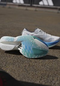 Witte hardloopschoenen met blauwgroene patroonzolen die op een gestructureerde buitenoppervlakte liggen, één schoen toont het merk "ADIZERO".