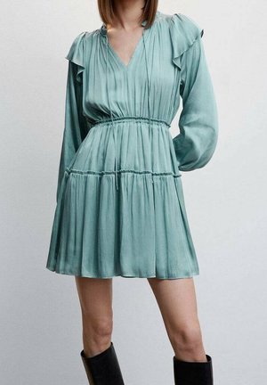 Femme portant une mini-robe à manches longues bleu clair avec une taille froncée et des détails volants aux épaules, assortie à des bottes noires montantes jusqu'aux genoux.