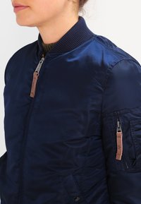 Chaqueta bomber de color navy con acabado satinado, cuello acanalado, bolsillos laterales y un bolsillo adicional con cremallera en la manga, con detalles de herrajes en color marrón.