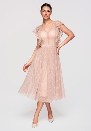 Kobieta stojąca w prześwitującej sukience w groszki do kolan z falbankowymi rękawami, nosząca szpilki z ostrym czubkiem i pozująca z jedną ręką na biodrze.