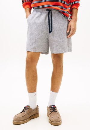 Άνδρας που φοράει ανοιχτό γκρι σορτς με κορδόνι, λευκές κάλτσες μέχρι τον αστράγαλο, καφέ δερμάτινα παπούτσια με κορδόνια και πορτοκαλί μακρυμάνικο πουκάμισο με ρίγες, με τα χέρια στις τσέπες.