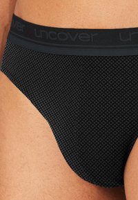 Sort undertøj med et struktureret mønster, der har en bred elastisk linning prægede med "uncover." Nærbillede af sidesømmen.