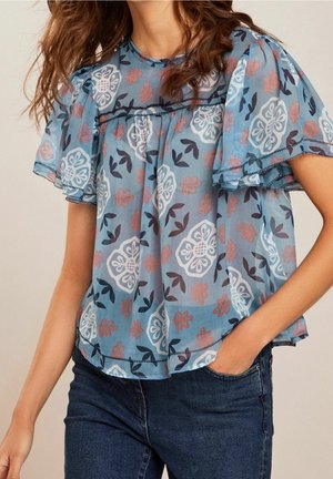 Femme portant un chemisier bleu transparent à motifs floraux avec des manches courtes volantées et un jean bleu foncé, main dans la poche, sur un fond neutre.