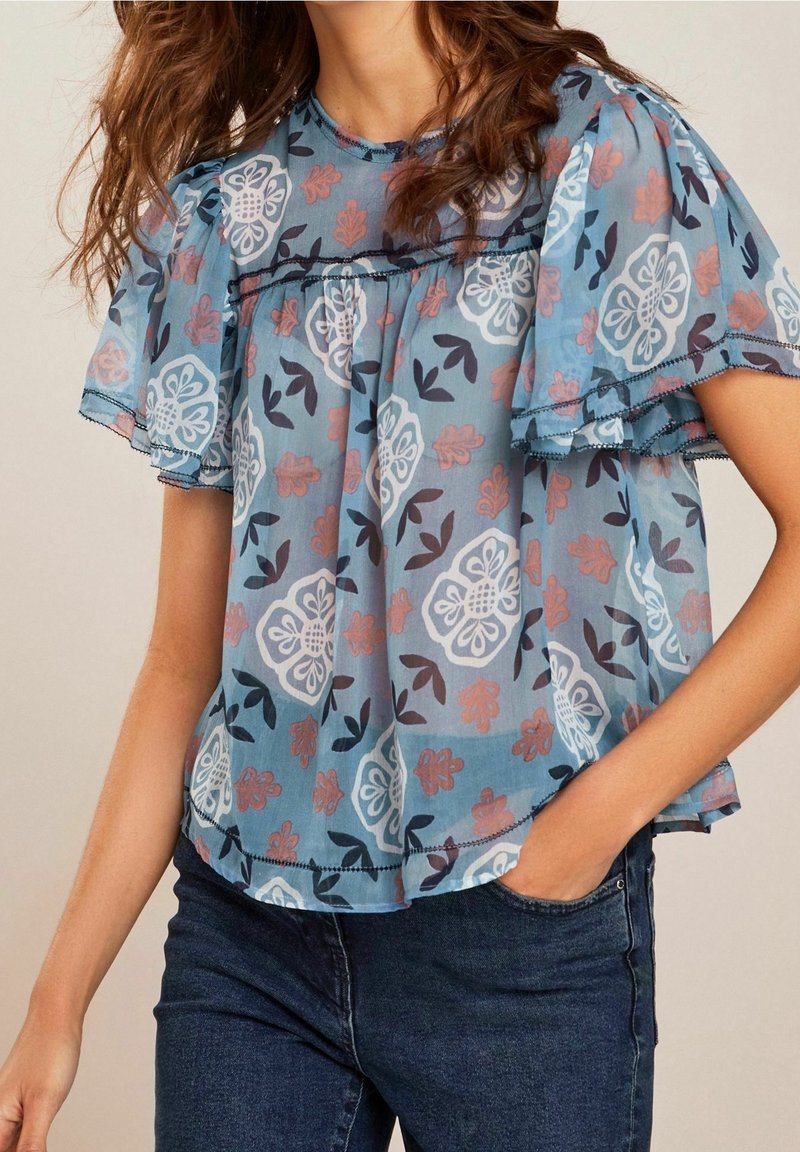 Femme portant un chemisier bleu transparent à motifs floraux avec des manches courtes volantées et un jean bleu foncé, main dans la poche, sur un fond neutre.