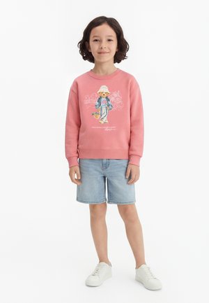 Copil cu păr maro până la umeri poartă un hanorac roz cu un desen cu ursuleț, pantaloni scurți de denim deschis și adidași albi, stând în fața unui fundal alb.