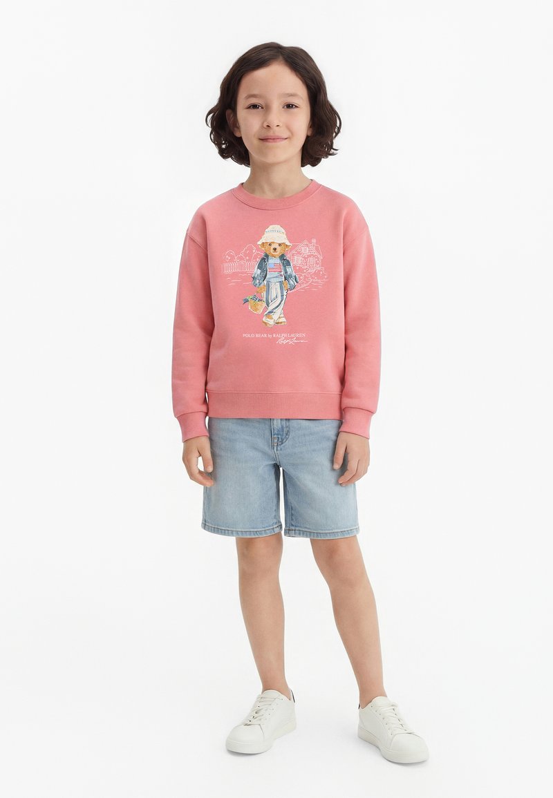 Παιδί με καστανά μαλλιά μέχρι τους ώμους φοράει ροζ φούτερ με σχέδιο αρκούδας, ανοιχτόχρωμα τζιν σορτς και λευκά αθλητικά παπούτσια, στέκεται μπροστά σε λευκό φόντο.