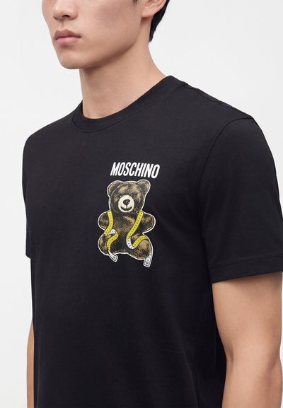 Νέος άνδρας φοράει μαύρο t-shirt Moschino με εκτύπωση αρκουδάκι τυλιγμένο σε κίτρινη μεζούρα στο στήθος.