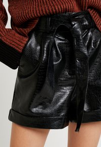 Černé šortky z imitace kůže s krokodýlím vzorem, opatřené uvázaným páskem a ohrnutými manžetami. Kapsy jsou integrované do designu.