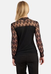 Blusa nera con lunghe maniche in pizzo trasparente, caratterizzata da motivi floreali. Il corpo principale è di un nero uniforme, con una texture liscia e un design aderente.
