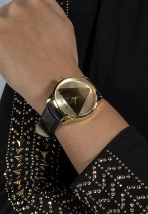 Montre en or avec un cadran triangulaire, bracelet en cuir noir et accents dorés. La robe possède des clous métalliques et une silhouette ajustée.
