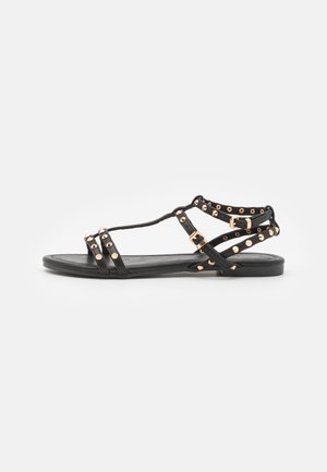 Sort flad sandal med flere remme, guldfarvede nitter og justerbare spænder på en ensartet hvid baggrund.