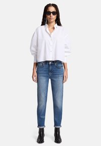 Camicia bianca cropped con bottoni e maniche a palloncino, abbinata a jeans blu a gamba dritta e stivaletti neri. Gli occhiali da sole neri aggiungono contrasto.