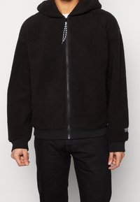 Mann trägt einen schwarzen Fleece-Hoodie mit Reißverschluss und Kordelzügen sowie schwarze Hosen, steht vor einem schlichten Hintergrund, Hände entspannt an den Seiten.