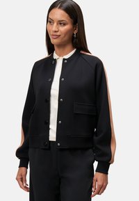 Black bomber jacket with contrasting light brown shoulder accents, snap buttons, ribbed cuffs, and a front pocket. Soft fabric texture.