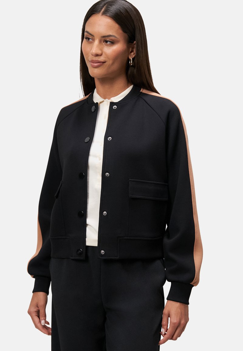 Black bomber jacket with contrasting light brown shoulder accents, snap buttons, ribbed cuffs, and a front pocket. Soft fabric texture.