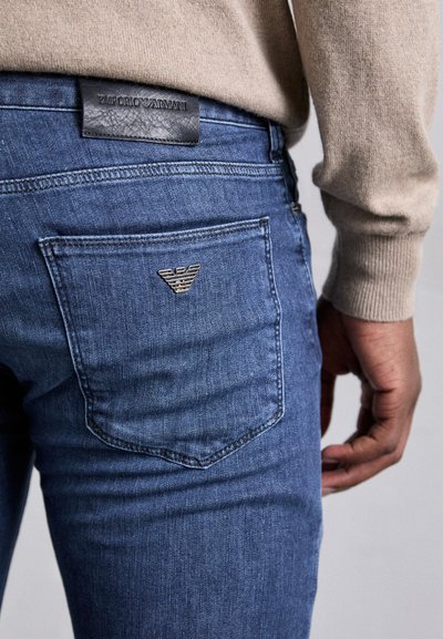 Jeanși din denim albastru cu o etichetă din piele neagră texturată. Prezintă un mic logo metalic pe buzunarul din spate. Design ajustat și decolorare subtilă.