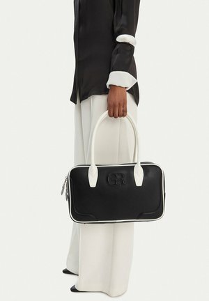 Persona con blusa negra y pantalones blancos sosteniendo un bolso rectangular negro con asas blancas y el logo en relieve "GR".