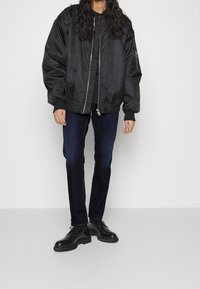 Černá bomber bunda se zipem, žebrovanými manžetami a bočními kapsami, spárovaná s tmavými džíny a černými šn�ěrovacími botami.