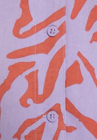 Kortärmad skjorta i ljuslila tyg med ett djärvt orange abstrakt mönster; har en knappslutning med en liten rund lila knapp.