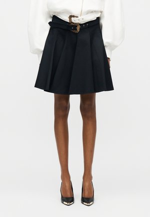 Femme portant une jupe noire plissée avec une large ceinture ornée d'une grande boucle élaborée, associée à des escarpins noirs à bout pointu et un chemisier blanc.