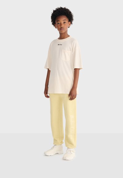 Ung person med krøllet hår iført en oversized cremefarvet T-shirt og gule joggingbukser, stående foran en ensfarvet baggrund.