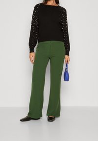 Schwarzer Pullover mit Perlenverzierungen an den Ärmeln, kombiniert mit grünen, weit geschnittenen Hosen. Dazu eine kleine blaue Handtasche und schwarze Ballerinas.