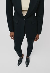 Musta muotoiltu bleiseri terävillä läpillä, yhdessä räätälöityjen mustien housujen ja kärkitoisilla mustilla korkokengillä, vaaleanbeige topin päällä.