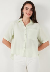 Camicia button-up a maniche corte verde chiaro con taschino sul petto, realizzata in tessuto leggero, caratterizzata da un orlo dritto e maniche arrotolate.