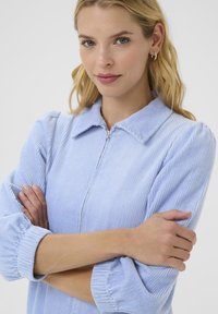 Una donna con lunghi capelli biondi posa con le braccia incrociate, indossando un top in corda azzurro chiaro con zip, sorridendo dolcemente. Sfondo neutro.