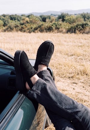 Une personne chaussée de chaussures noires sans lacets pose ses jambes croisées sur la portière d’une voiture, près d’un champ sec et de collines bordées d’arbres.