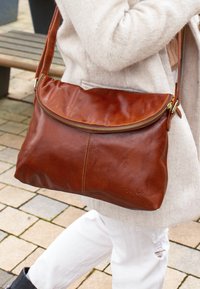 Borsa a spalla in pelle marrone con chiusura zip, texture morbida e forma arrotondata. Presenta dettagli di cucitura e una tracolla comoda.