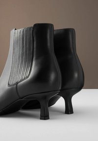 Černé kožené kotníkové boty s špičatou špičkou, žebrovanými detaily po stranách a štíhlým, zakřiveným podpatkem. Hladký povrch po celém obvodu.