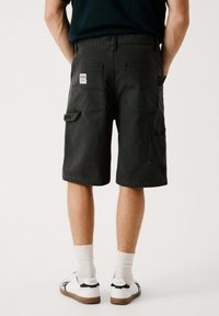 Pantalones cortos cargo negros con múltiples bolsillos, un corte relajado y una textura suave. Llevados con zapatillas blancas y calcetines de longitud media.