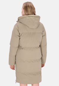 Vrouw staat met de rug zichtbaar, gekleed in een beige gewatteerde winterjas met capuchon tegen een effen lichte achtergrond.
