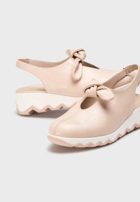 Zapatos slingback de cuero beige con un detalle de nudo en la parte frontal y una suela blanca gruesa con zigzagueo. Diseño de punta redondeada y parte trasera abierta.