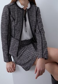 Tweed jacket in black, white, and purple with a zip closure and leather accents paired with a matching mini skirt. White blouse with frills.