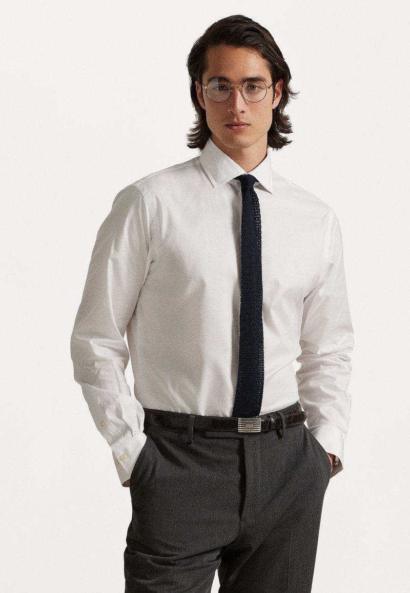 Valge särk krae ja tumeda tekstuuriga lipsuga, hallid püksid ning tume nahkrihm metallist sulemehega.