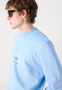 Felpa azzurra con scollo rotondo a costine, caratterizzata da un logo ricamato bianco sul davanti. Tessuto di cotone liscio con una vestibilità comoda.