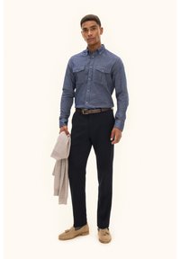 Camicetta di jeans button-up blu scuro con due tasche sul petto, abbinata a pantaloni navy su misura. Il modello tiene un maglione beige. Scarpe beige.