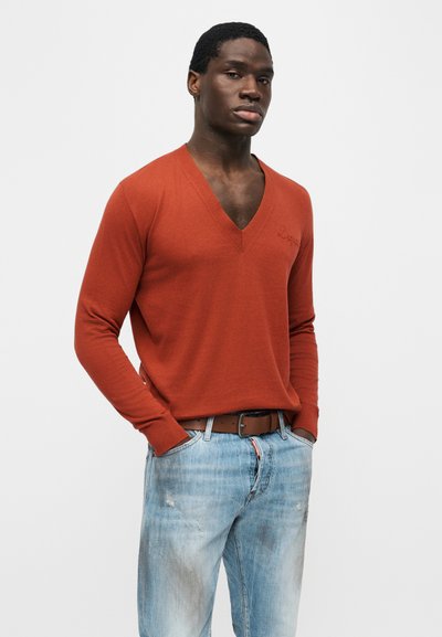 Jeune homme portant un pull col en V orange brûlé rentré dans un jean délavé bleu clair avec une ceinture marron, les mains dans les poches.