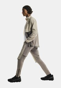 Giacca leggera beige con pannelli grigi e tasche con zip, abbinata a pantaloni tapered coordinati. Sneakers nere con suole texture.