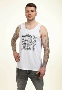 Top sin mangas blanco con una impresión gráfica negra de Mickey Mouse y sus amigos. El modelo tiene tatuajes y lleva puestos unos jeans grises desgastados.