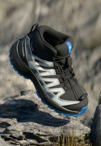 Juodas žygio batų modelis su pilkais akcentais, tekstūruota viršutine dalimi, mėlynais akcentais ir tvirtu guminio pado. Yra saugus raištelių sistemos funkcionalumas.