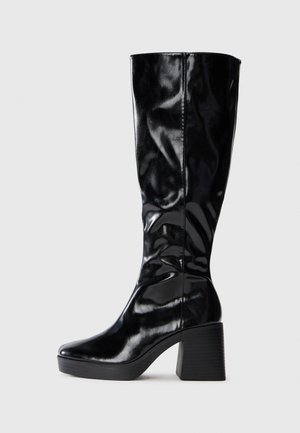 Botte noire, montée jusqu'au genou, en cuir verni brillant ; dotée d'un talon carré structuré et d'un bout arrondi. Design simpliste et élégant.