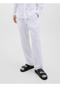 Camisa y pantalones de lino blancos, combinados con sandalias negras con correas y suela redondeada, sobre un fondo blanco liso.