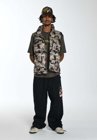 Kamuflażowa kamizelka puchowa z wysokim kołnierzem, zestawiona z ciemnozieloną koszulką i luźnymi czarnymi spodniami z haftowanym wzorem. Brązowa czapka z grafiką.