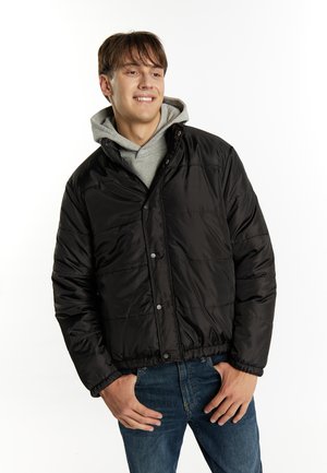 Sort pufferjakke med kviltet tekstur, trykknapper og elastiske manchetter, båret over en grå hættetrøje. Parret med blå jeans for et afslappet look.