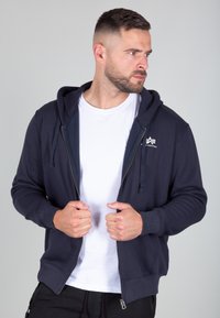 Felpa con zip di colore blu navy, con lacci, polsini a costine e tasca. Indossata sopra una t-shirt bianca, che mostra un design semplice e casual.