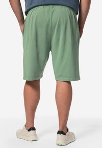 Pantaloni corti verdi in cotone con elastico in vita, lunghi fino al ginocchio, caratterizzati da una consistenza morbida. Indossati con sneakers leggere, che evidenziano un design casual.