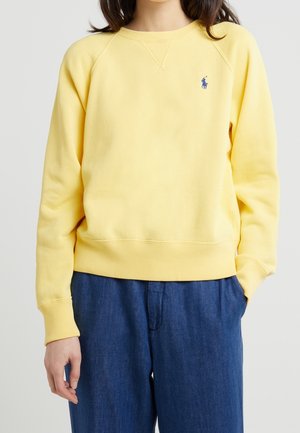 Personne portant un sweat-shirt à manches longues jaune clair avec un logo bleu brodé sur la poitrine et un pantalon bleu ample, debout devant un arrière-plan neutre.